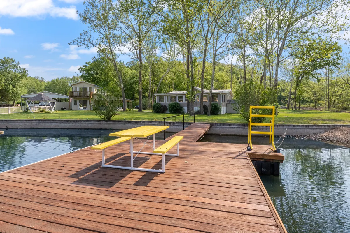 A yellow picnic table is positioned near the dock, with serene lake waters and trees in the background. The setting is ideal for sunset picnics, morning coffee, fishing, or launching small watercraft. This lively, inviting scene emphasizes nature and lakeside leisure.