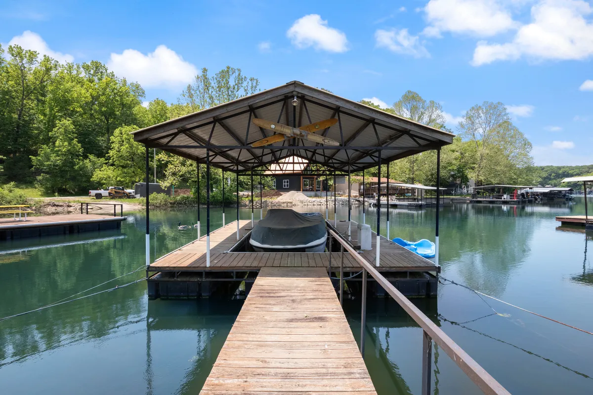 This image captures a serene lakeside scene with a boathouse, a dock extending over calm water, and lush surrounding trees. The area invites boating, fishing, and relaxation by the water, with ample space for social gatherings and enjoying the outdoors.