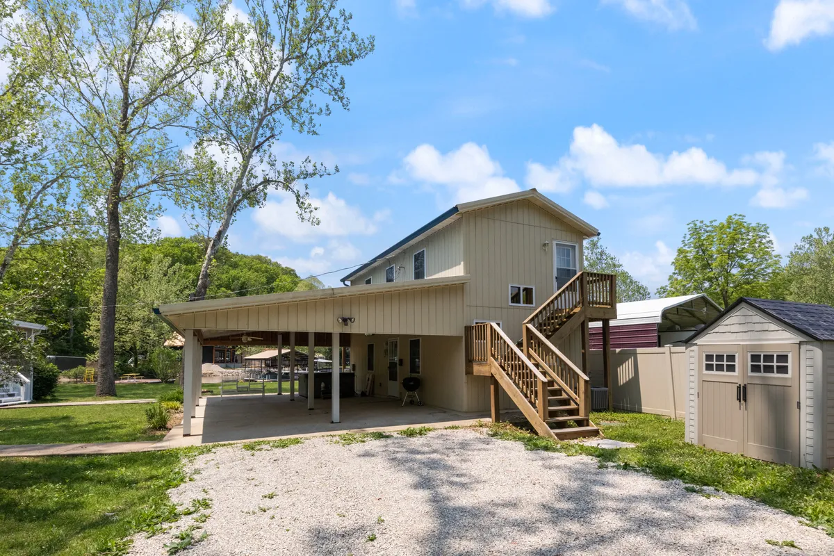 This inviting exterior photo features the house framed by mature trees, a staircase, a porch, and a garage shed. The setting is serene and perfect for summer parties, morning coffee under the trees, or enjoying sunsets from the porch.