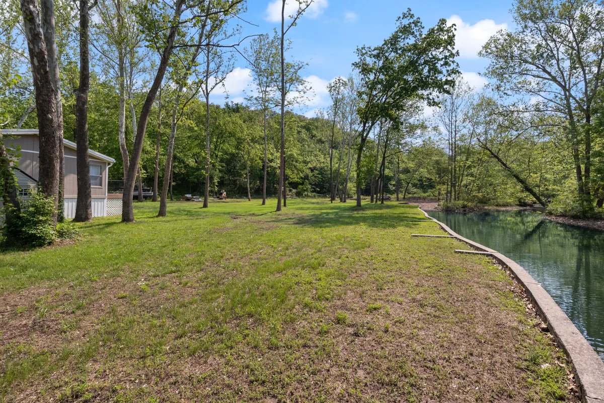 A lush, green lawn stretches down to calm water, framed by tall trees and a house visible in the background. The tranquil setting evokes a sense of escape and relaxation, perfect for enjoying nature, gathering for picnics or fireside chats, and watching the sunset by the lake.