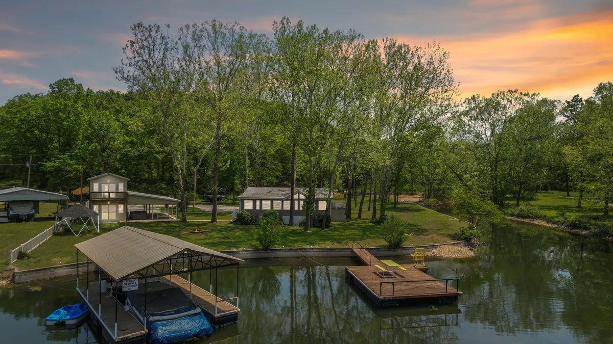 A wide view of the lakefront with a well-maintained dock stretching out over the water, flanked by two homes partially visible among mature trees. The sun is setting, creating a warm glow over the lake and inviting ambiance. The surrounding lawn offers plenty of space for outdoor activities.