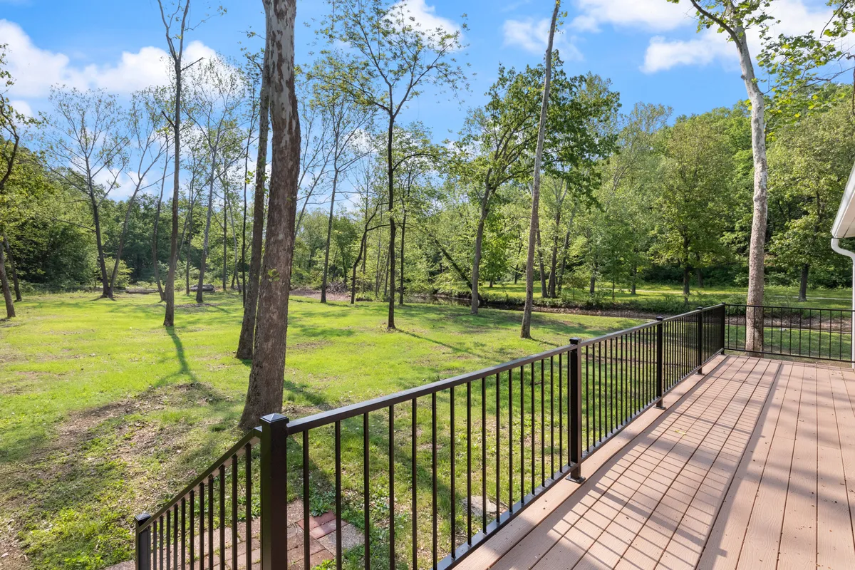 This exterior image shows a deck railing overlooking lush green grass, tall trees, and a clear blue sky. The peaceful setting is perfect for observing nature, relaxing outdoors, hosting family gatherings, or simply enjoying a book in the fresh air.