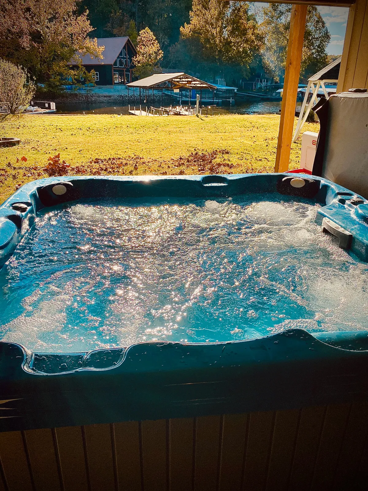The image showcases a serene lakeside setting at sunset, with a bubbling hot tub overlooking calm water, a private dock extending from a lush, expansive yard, and a second cozy home visible in the distance. The tranquil scene invites relaxation, with hints of activities like boating, fishing, or simply enjoying the view from the hot tub.
