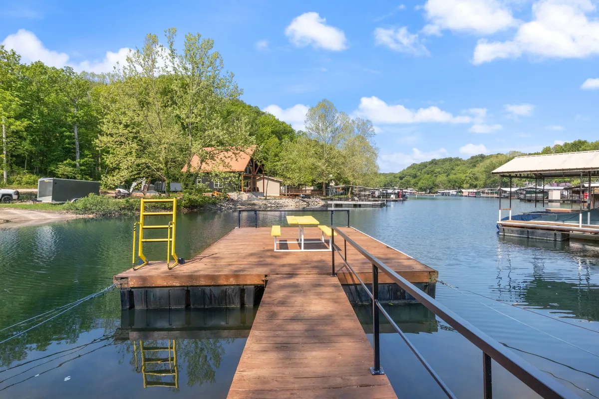 This image showcases a private dock extending over a tranquil lake, surrounded by lush trees and nearby boathouses. The calm water reflects the evening sky, and the dock invites activities like fishing, relaxing, or launching a kayak. The scene evokes a sense of peaceful lakeside living.