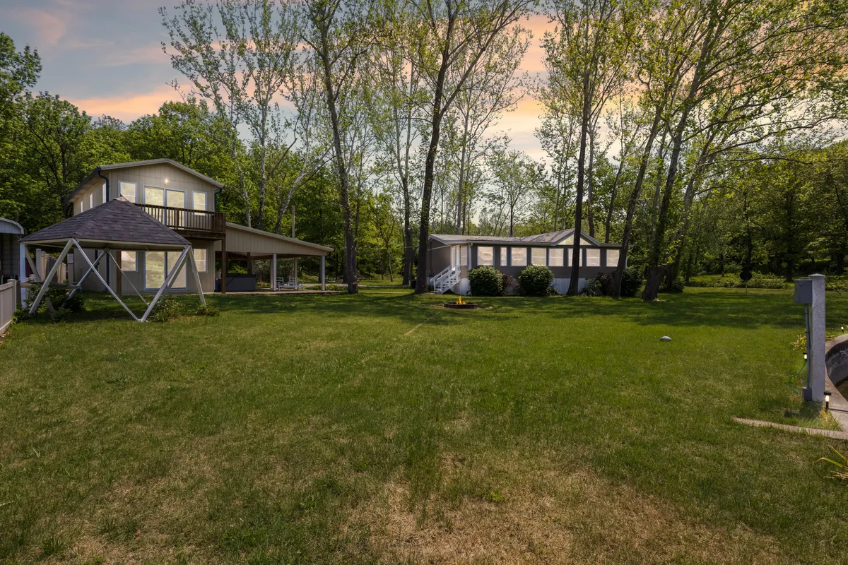 Two distinct buildings are nestled among mature trees, separated by an open lawn. The scene suggests space and privacy between the two homes, inviting guests to enjoy both shared and individual experiences within a serene, wooded environment.