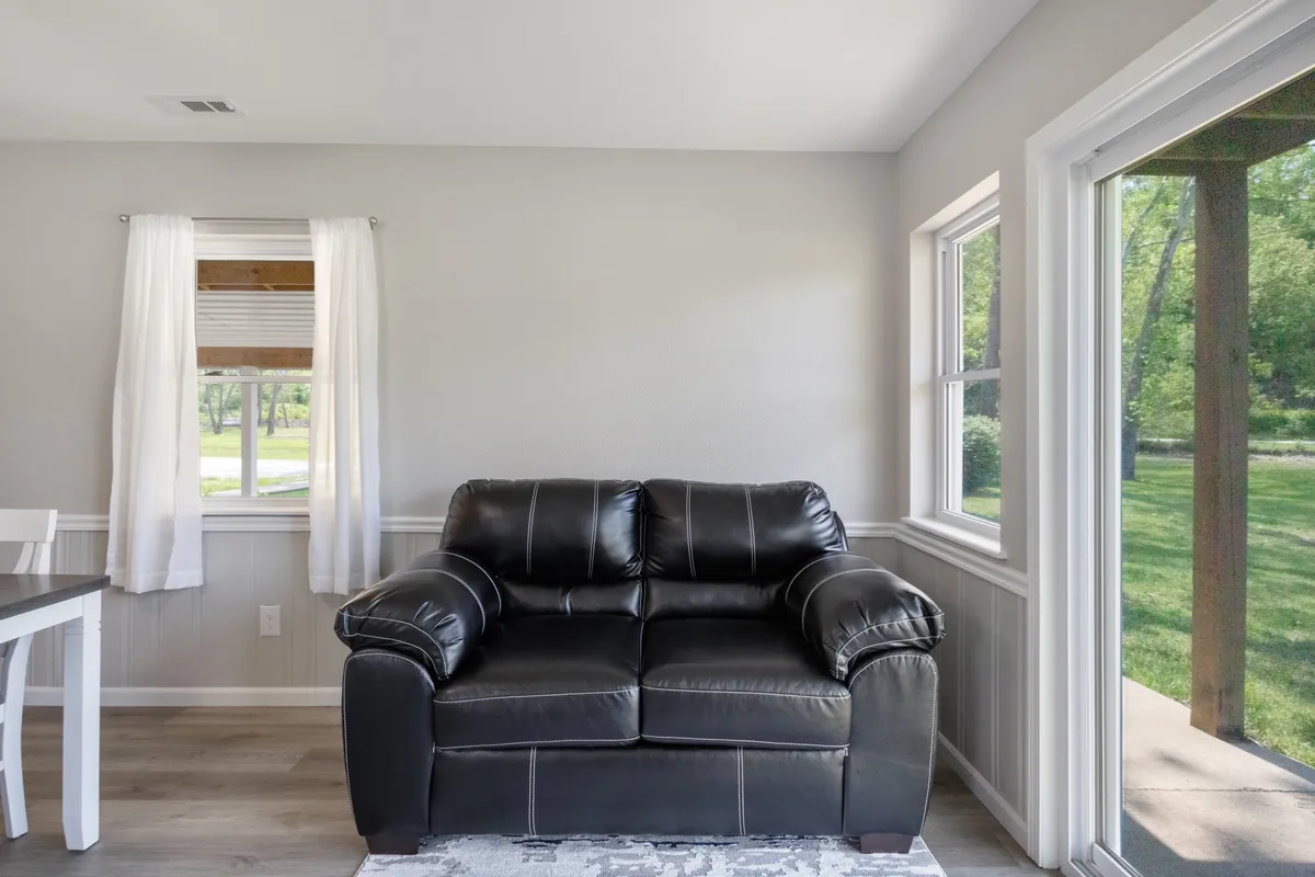 A spacious, light-filled living area featuring a black leather couch, large windows with white curtains, and a dining table. The room feels welcoming and organized, with ample space for relaxing or socializing. The clean, uncluttered look highlights the comfort and care taken in preparing the home for guests.
