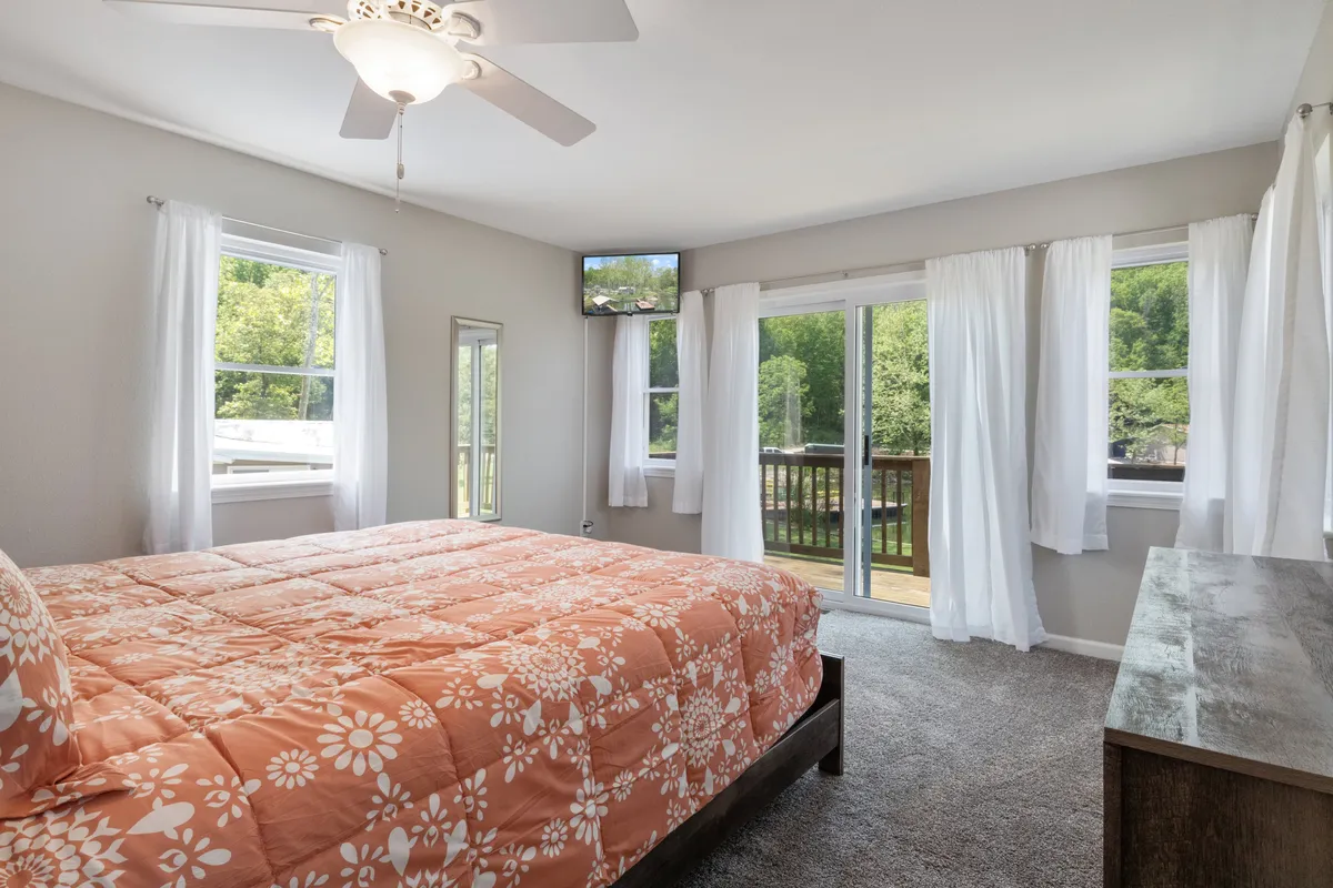 A cozy bedroom with a floral quilt on the bed, large windows, a sliding glass door leading outside, a ceiling fan, and views of nature. The space feels private and relaxing—ideal for downtime and personal moments.