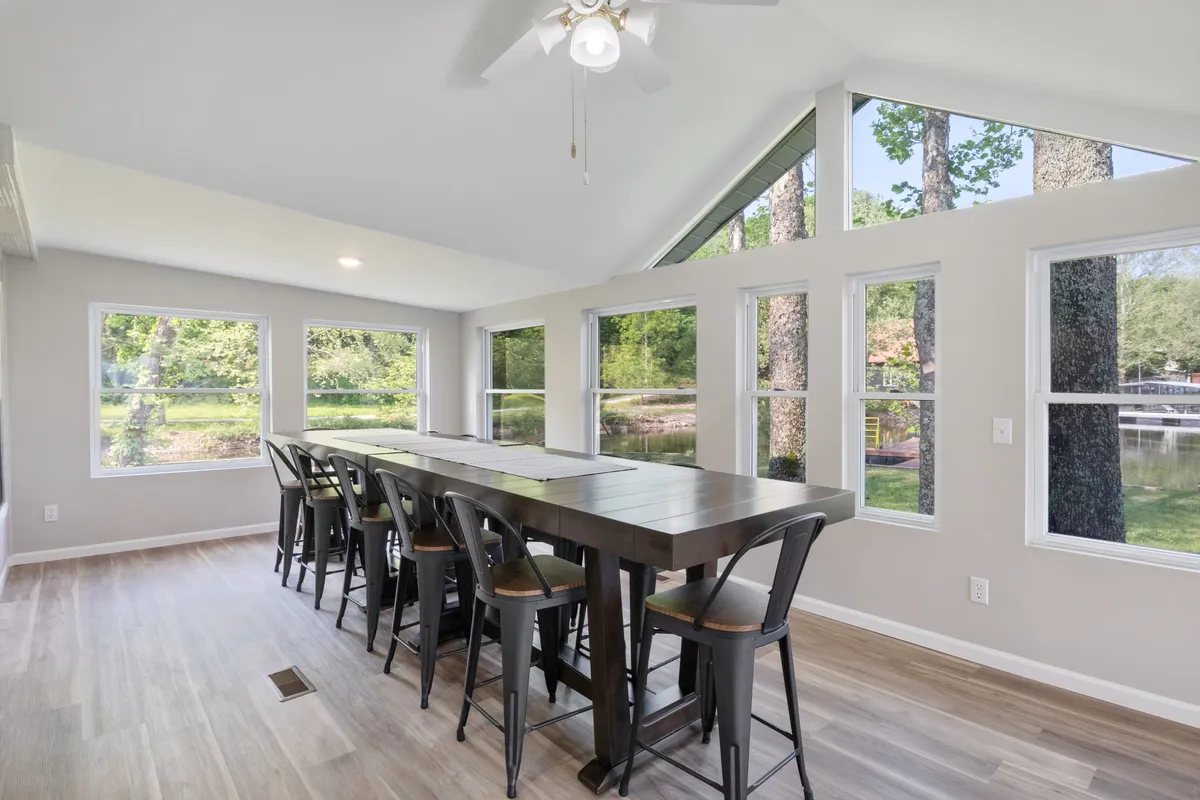 A large dining table sits by expansive windows overlooking the natural landscape, filling the room with natural light. The setup suggests space for group meals, games, or celebrations.